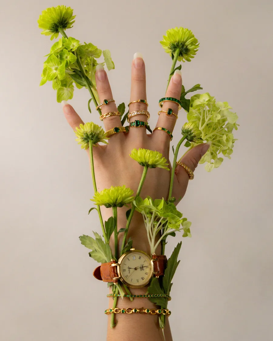 A raised hand wearing rings, bracelets and a watch, threaded with green flowers