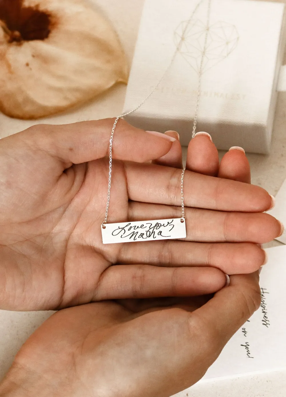 Open hands holding a silver bar necklace engraved with a handwritten message, the gift box behind