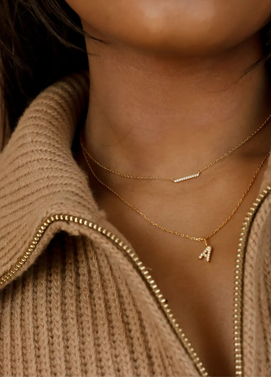 A bar necklace layered over an initial A necklace, worn with a knit collar