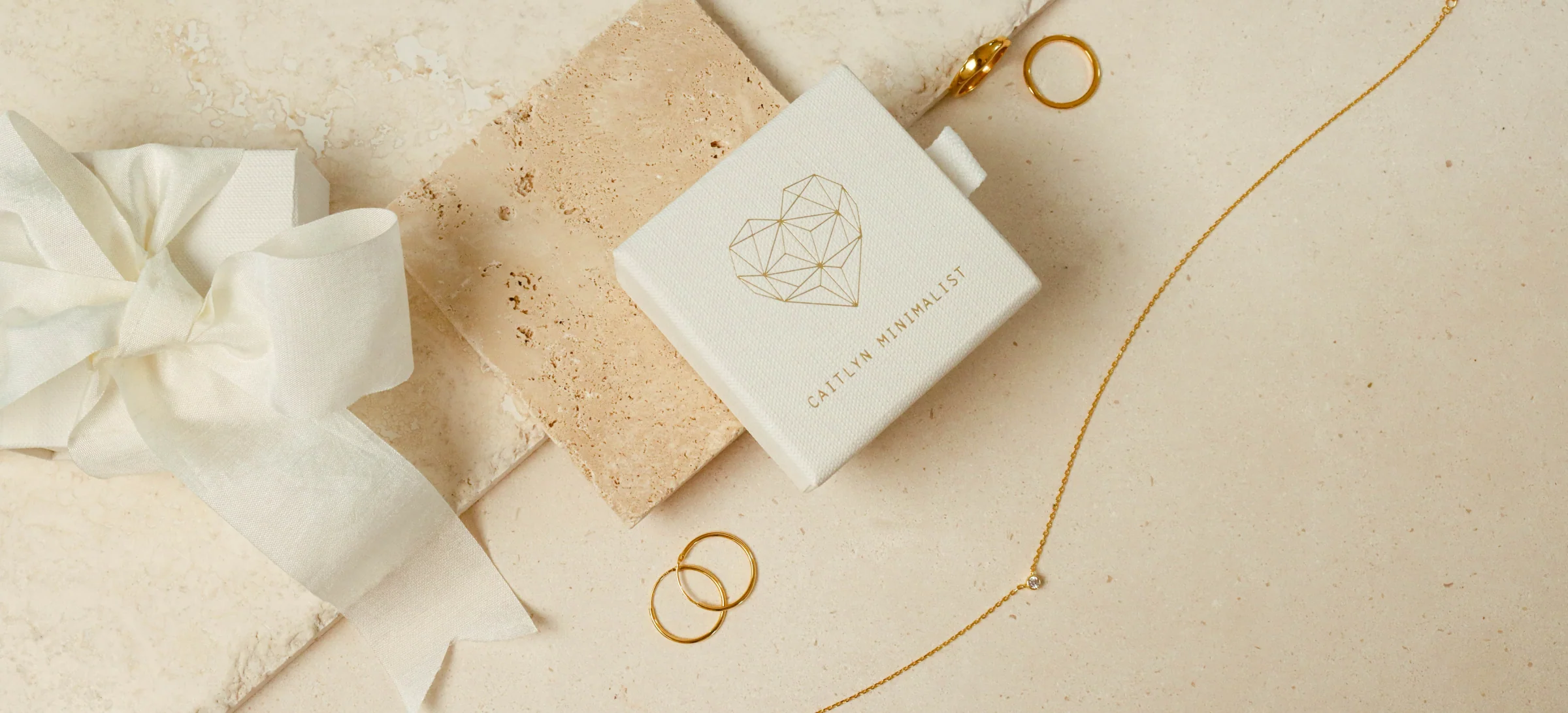 A Caitlyn Minimalist gift box with the heart logo beside a ribbon bow, rings and a fine chain