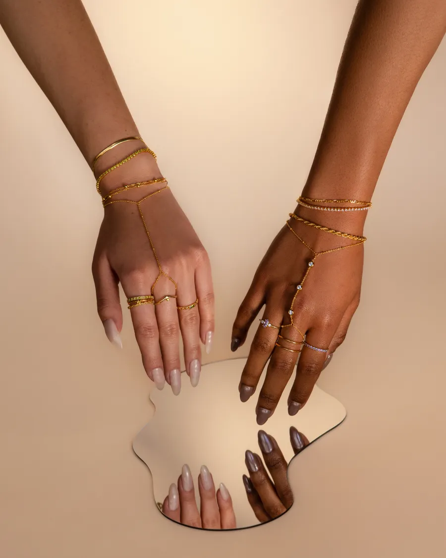 Two hands wearing fine gold rings and bracelets, resting on a small mirror